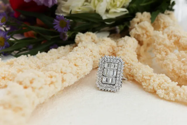 A baguette diamond cluster ring with a double halo in a taupe velvet box among cream flowers