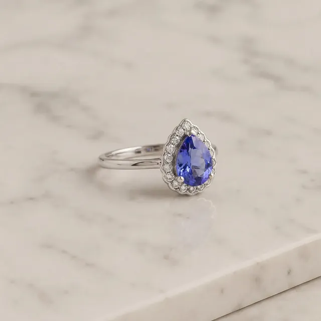 A white gold ring with a blue pear stone in a diamond halo on marble