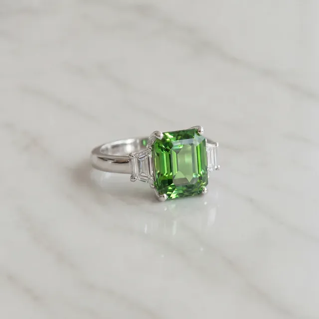A white gold ring with a green emerald cut stone on marble